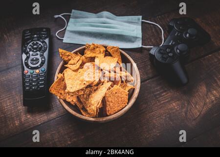 Télécommande, console de jeux rc, bol de chips et masque médical sur table en bois. Coronavirus Covid-19 quarantaine rester à la maison concept. Banque D'Images