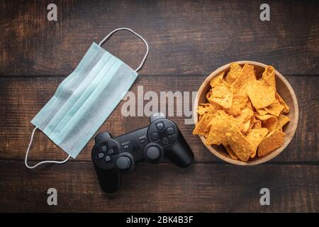 Console de jeu rc, bol de chips et masque médical sur table en bois. Coronavirus Covid-19 quarantaine rester à la maison concept. Banque D'Images