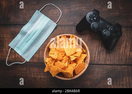 Console de jeu rc, bol de chips et masque médical sur table en bois. Coronavirus Covid-19 quarantaine rester à la maison concept. Banque D'Images