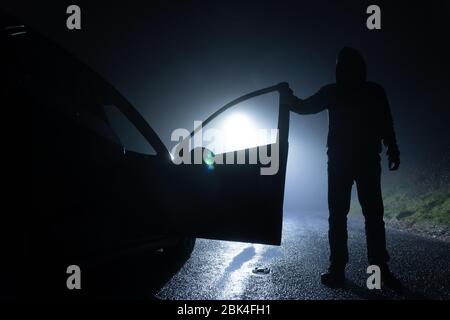 Un homme debout à côté d'une voiture, avec la porte ouverte, sous un feu de rue. Une nuit d'hiver brumeuse Banque D'Images