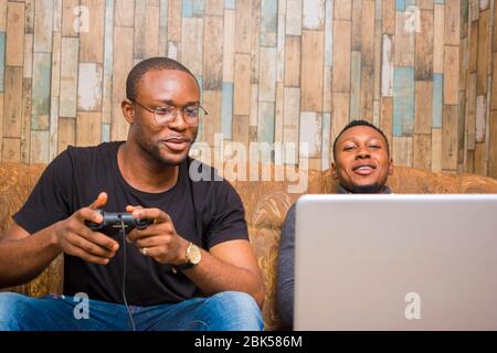 deux gars africains de belle jouants assis sur le canapé jouant aux jeux vidéo avec joystick, manette de jeu, pad Banque D'Images