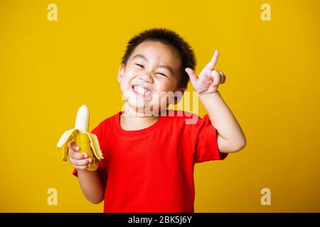 Joyeux portrait enfant ou enfant asiatique mignon petit garçon beau sourire portant un t-shirt rouge jouant tient pelé banane pour manger, studio tourné isolé sur Banque D'Images