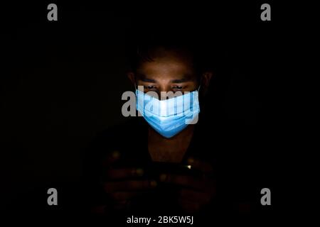 Un jeune homme bleu portant un masque chirurgical jouait à Dhaka pour jouer à des jeux sur smartphone. Banque D'Images