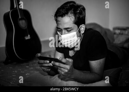 Un jeune homme portant un masque chirurgical jouait à des jeux mobiles chez lui en raison de la quarantaine à domicile du coronavirus à Dhaka. Banque D'Images