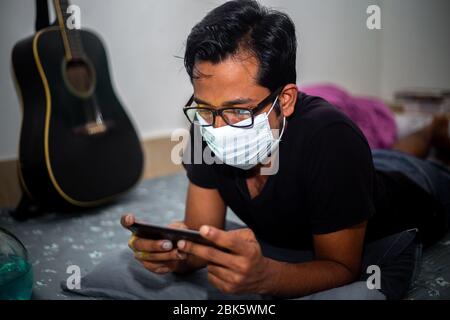 Un jeune homme portant un masque chirurgical jouait à des jeux mobiles chez lui en raison de la quarantaine à domicile du coronavirus à Dhaka. Banque D'Images