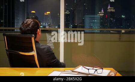 Homme d'affaires asiatique regardant à l'extérieur de la fenêtre tout en examinant un bref document après le travail. Salle de réunion située à l'étage supérieur, la nuit. Bangkok est très animée Banque D'Images