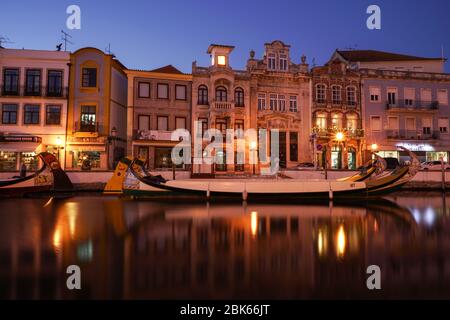 Aveiro, Portugal - avril 2019 : longue exposition des bateaux Moliceiro dans le canal de l'eau avec des bâtiments Art nouveau, après le coucher du soleil. Banque D'Images