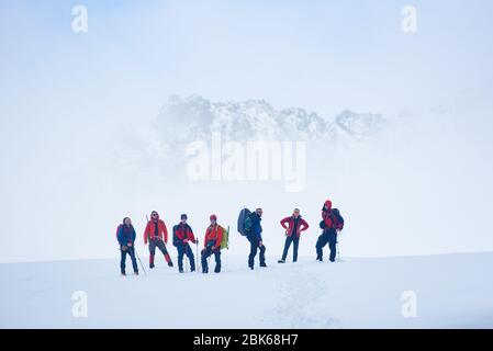 Équipe de randonneurs debout sur une vallée de flanc de colline recouverte de neige. Alpinistes et bâtons de randonnée mâles profitant de la vue sur les montagnes d'hiver tout en prenant repos pendant la randonnée, regardant l'appareil photo Banque D'Images