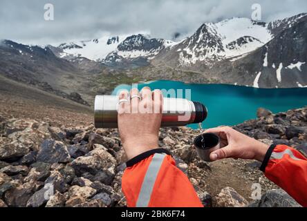 Verser le thé chaud de l'homme à la Tasse thermos au lac de montagne l'arrière-plan. Le Lac Karakol Ala-Kul national park, le Kirghizistan Banque D'Images