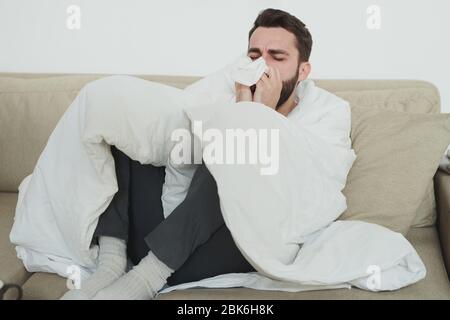 Jeune homme avec grippe ou covid19 toussant dans le mouchoir ou le tissu en papier tout en assis sur le canapé dans le salon pendant l'auto-isolation Banque D'Images
