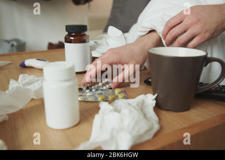 Mains de l'homme malade prenant des comprimés de table tout en assis sur le lit ou le canapé près de par et en choisissant des antipyrétiques parmi la variété de médicaments Banque D'Images