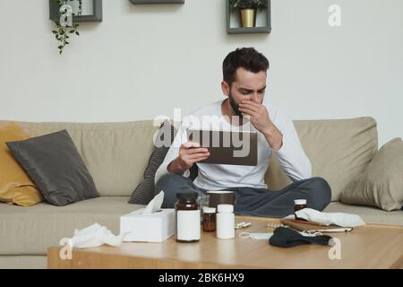 Jeune homme très malade avec tablette numérique assis sur le canapé et couvrant sa bouche tout en toussant pendant le travail à distance dans l'environnement de la maison Banque D'Images