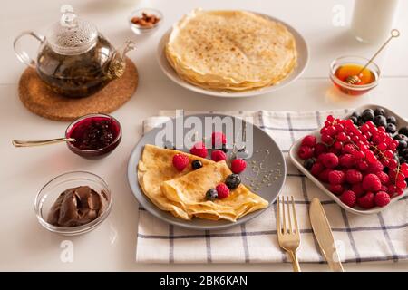 Table de cuisine pour le petit déjeuner avec des crêpes maison appétissantes sur plaque, baies fraîches, thé, miel, tartiner au chocolat et confiture de cerises Banque D'Images