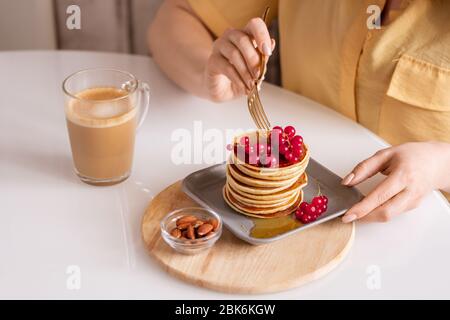 Les mains de la jeune femme affamée assis à la table et avoir de délicieuses crêpes au miel et au currant rouge frais avec cappuccino dans le café Banque D'Images