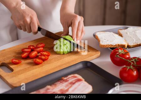 Mains de femme de ménage avec couteau coupant l'avocat frais et les tomates sur la planche en bois pour les sandwichs tout en cuisinant le petit déjeuner pour elle-même Banque D'Images