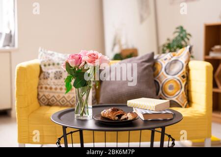 Deux livres, croissant frais sur plaque et bouquet de roses roses roses dans un verre d'eau sur une petite table avec canapé sur fond dans le salon Banque D'Images