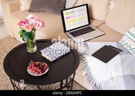 Raisins rouges sur soucoupe, roses roses dans le verre d'eau et réserver sur petite table par canapé avec ordinateur portable, coussin et stylet dans la chambre domestique Banque D'Images