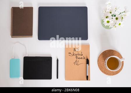 Disposition plate d'une tasse de thé vert, fleurs blanches dans la verseuse, croquis avec surligneur, un coussin avec stylet, un carnet et un ordinateur portable plié sur le bureau Banque D'Images