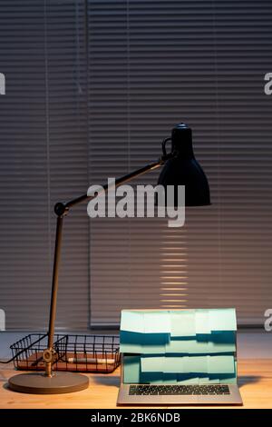 Lumière de la lampe sur l'ordinateur portable avec des notes bleues sur l'écran et panier avec surligneur près de la table en bois par fenêtre avec jalousie Banque D'Images