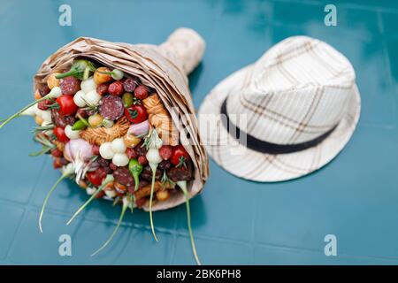 Le bouquet original composé de saucisses fumées, fromage, ail, tomates et poivrons se trouve sur une table en plastique près du chapeau de homme. Banque D'Images