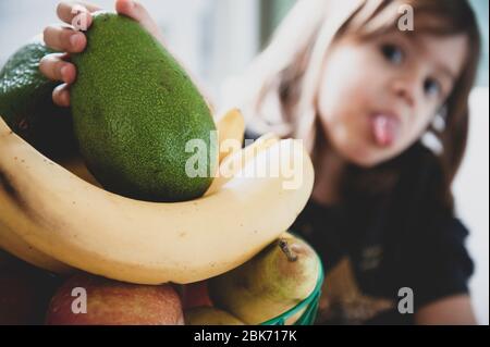 Petite fille prendre main un avocat à partir d'un panier de fruits. Main de l'enfant tenant vert frais mûr avocat. Nourriture saine pour les enfants. Face drôle de petit rif Banque D'Images