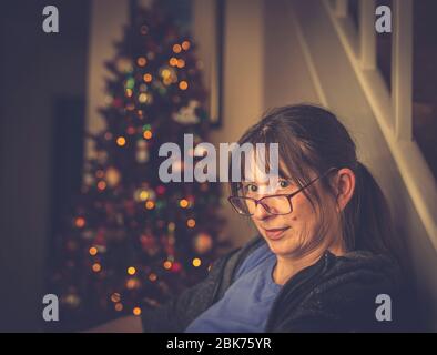Gros plan d'une femme fatiguée assise sur une chaise, regardant par-dessus le dessus de ses verres, se détendant par un arbre de Noël britannique décoré. Banque D'Images
