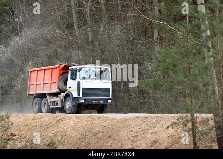 Camion lourd de plusieurs tonnes avec cabine blanche et corps orange se déplace le long de la route poussiéreuse de campagne dans avec fond de forêt de printemps, laissant derrière les nuages de d Banque D'Images