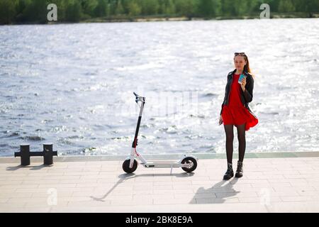 La jeune blonde se tient près du scooter électrique blanc sur la promenade qui tient du café Banque D'Images