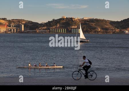 Homme vélo sur le bord du Tage avec équipe d'aviron et bateau à voile en arrière-plan, Lisbonne, Portugal Banque D'Images