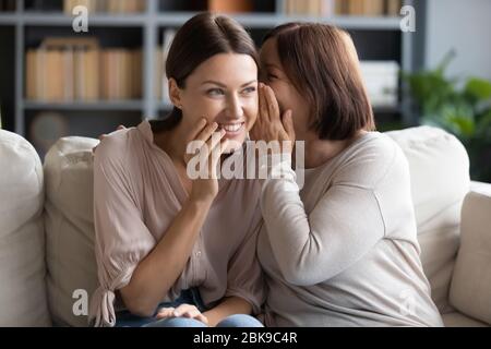 Mère d'âge moyen chuchotant dans l'oreille souriante de fille adulte, le commérages Banque D'Images