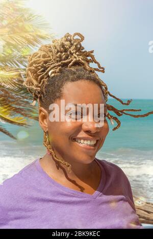 Gros plan Portrait de la belle femme noire, africaine ou caribéenne qui rit à l'extérieur et qui surplombe la magnifique plage et l'océan. Banque D'Images