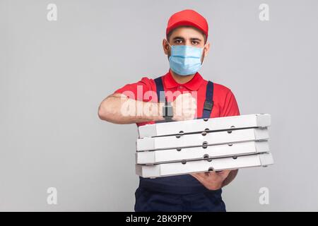 Dans les délais de livraison en quarantaine ! homme avec masque médical chirurgical en uniforme et rouge t-shirt debout, tenant pile de boîtes à pizza en carton sur bac gris Banque D'Images