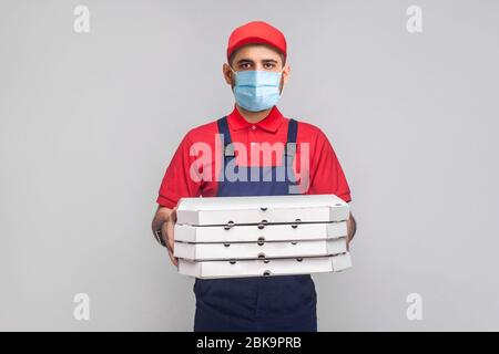 Livraison en quarantaine. Jeune homme avec masque médical chirurgical en uniforme bleu et t-shirt rouge debout et tenant pile de boîtes à pizza en carton sur gr Banque D'Images