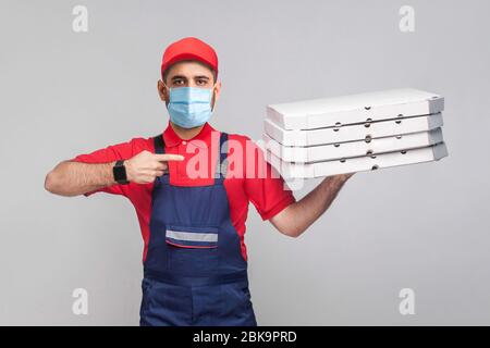 Livraison de pizza en quarantaine. Jeune homme avec masque médical chirurgical en uniforme bleu et t-shirt rouge debout, tenant et pointant le doigt à la pile de c Banque D'Images