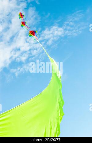 Cerfs-volants multicolores volant haut à l'extrémité d'un ruban vert vif contre un ciel bleu d'été et des nuages clairs Banque D'Images