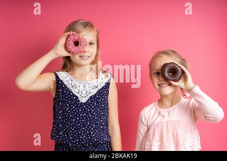 Deux filles blondes vêtues de robes colorées. Ils sourient et regardent à travers les trous de beignets dans leurs mains. Pose sur fond rose Banque D'Images