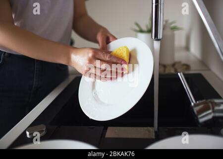 Jeune femme en cuisine pendant la quarantaine. Plaques blanches avec éponge et lave-vaisselle. Nettoyage de la vaisselle seul dans la cuisine. Vue de coupe. Banque D'Images