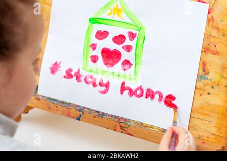 L'enfant écrit des mots rester maison avec l'aquarelle rouge sous la maison peinte avec des coeurs sur le chevalet. Concept de séjour à la maison. Banque D'Images