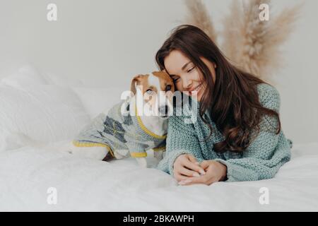 Photo de la belle femme brune en pull se trouve ensemble chien wih sur le lit mou, aime passer du temps avec l'animal préféré, se soucie des animaux, s Banque D'Images