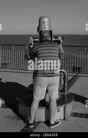 Homme regardant trop la mer à travers un télescope étrange depuis Southwold Pier suffok uk en noir et blanc Banque D'Images