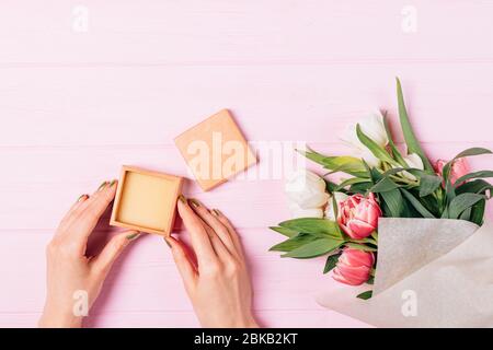 Vue de dessus des mains de la femme avec manucure verte tenant une petite boîte à cadeaux ouverte à côté du bouquet de fleurs fraîches sur table en bois rose. Banque D'Images