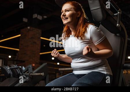 une fille en surpoids excitée montrant le geste du gagnant tout en étant assise sur une machine de fitness Banque D'Images