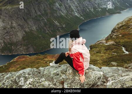 Fille dans une veste rose se trouve au bord d'une falaise sur fond des montagnes de Norvège et du ciel nuageux Banque D'Images