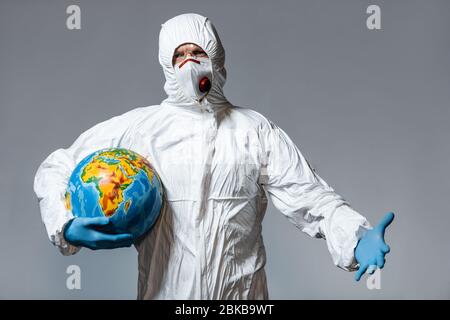 homme en costume de hazmat et masque médical tenant globe et gestuelle isolé sur gris Banque D'Images