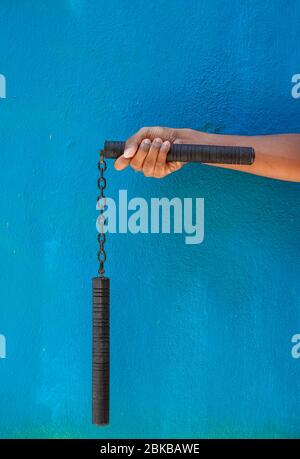 Garçon tenant Nunchaku sur sa main isolé sur fond bleu. Formation nunchaku noir pour débutant. Équipement d'arts martiaux Hot Tactical Gear. Banque D'Images