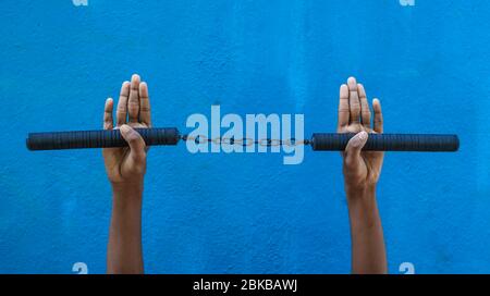 Garçon tenant Nunchaku sur sa main isolé sur fond bleu. Formation nunchaku noir pour débutant. Équipement d'arts martiaux Hot Tactical Gear. Banque D'Images
