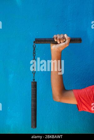 Garçon tenant Nunchaku sur sa main isolé sur fond bleu. Formation nunchaku noir pour débutant. Équipement d'arts martiaux Hot Tactical Gear. Banque D'Images