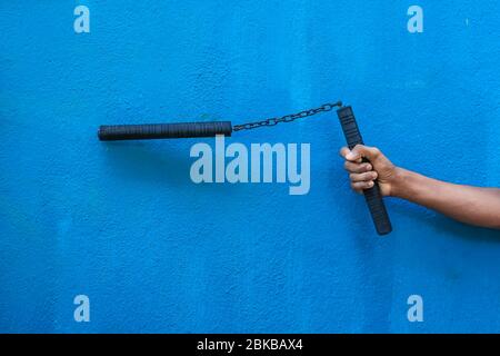 Garçon tenant Nunchaku sur sa main isolé sur fond bleu. Formation nunchaku noir pour débutant. Équipement d'arts martiaux Hot Tactical Gear. Banque D'Images