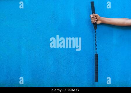 Garçon tenant Nunchaku sur sa main isolé sur fond bleu. Formation nunchaku noir pour débutant. Équipement d'arts martiaux Hot Tactical Gear. Banque D'Images