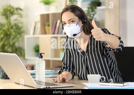 Bonne femme de télévendeur avec le pouce sur le confinement de covid-19 évitant la contagion avec le masque travaillant sur ordinateur portable à domicile Banque D'Images